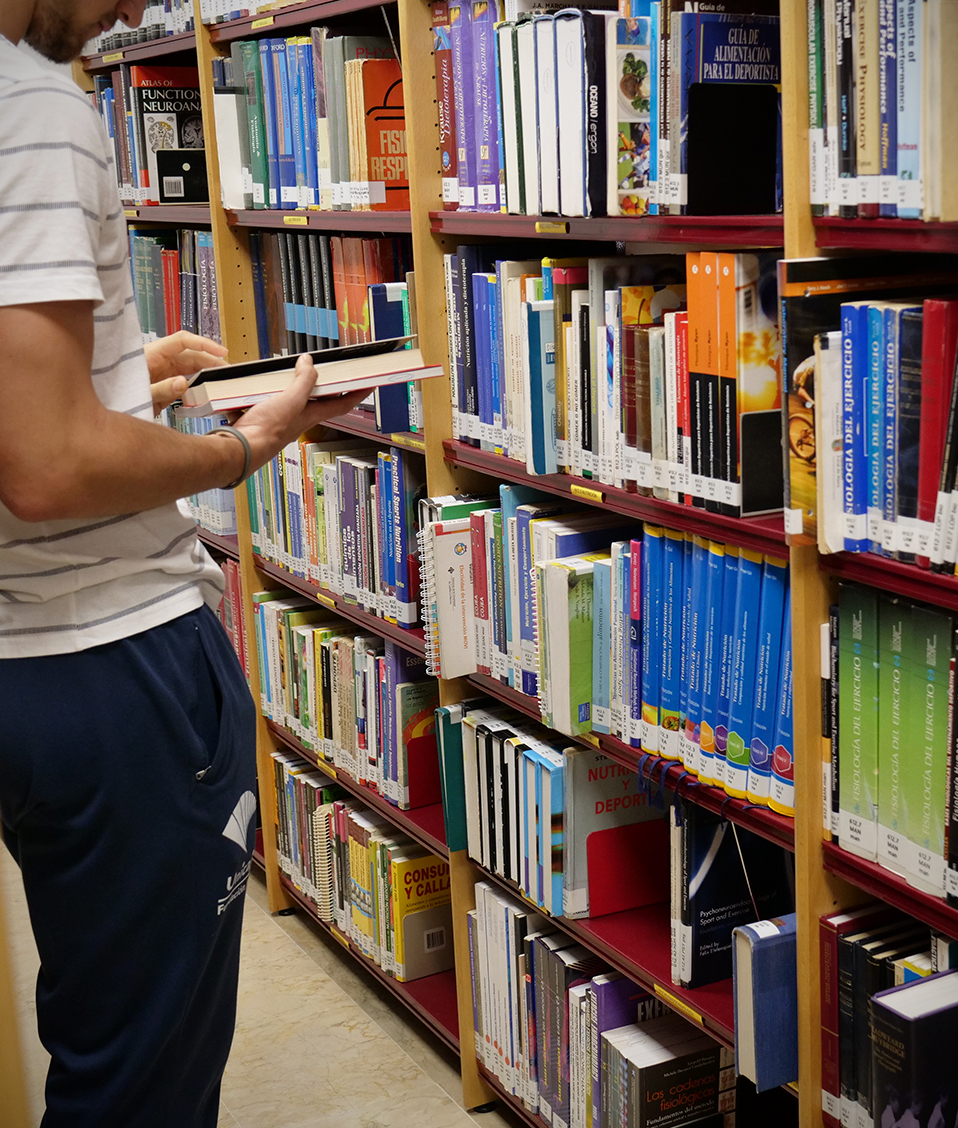 Un estudiante observa un libro frente a una estantería repleta de títulos en la biblioteca de la Facultad de Ciencias del Deporte.